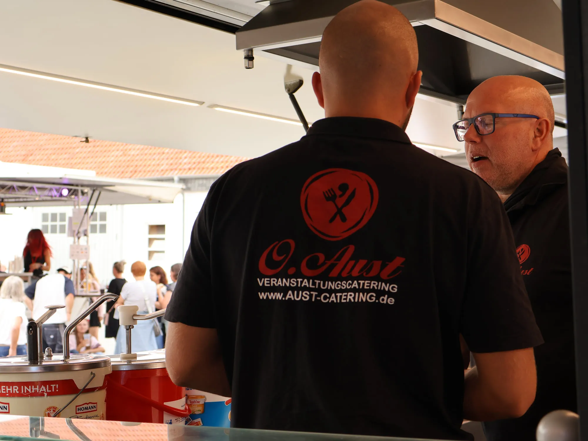 Zwei Mitarbeiter von Aust Catering im Cateringwagen, davor wartende Gäste